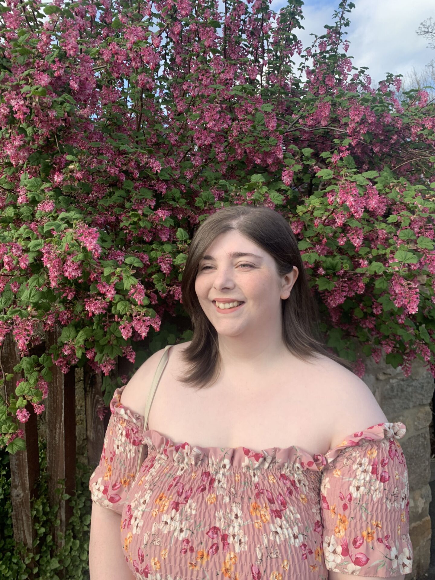 plus size brunette woman wearing a pink dress with white yellow and dark flowers on that stops at the shoulders and a neutral cream bag over her shoulder, standing in front of pink falling flowers.